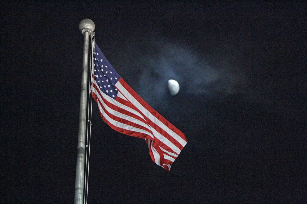An American flag on a pole at night against a dark sky with the moon behind